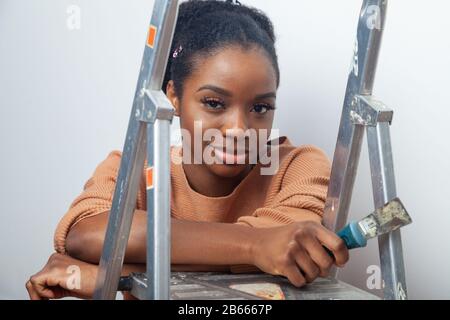 Eine schöne junge Frau afrikanischer Ethnie, die ihre Hände auf einer Stufenleiter ausruht und einen Tapetenkratzer in der Hand hält Stockfoto