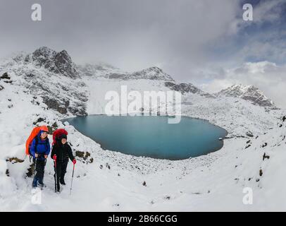 Zwei Wanderer fühlen sich klein vor dem riesigen blauen Bergsee in winterlichen verschneiten Bergen stehend Stockfoto