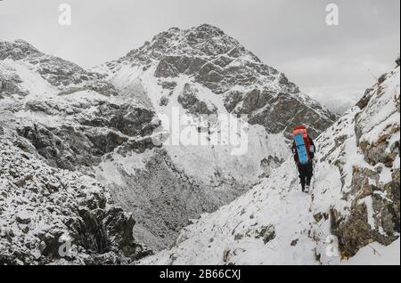 Kletterer, der auf einem steilen, schneebedeckten Hang in den Bergen auf dem Hintergrund einer Klippe und des Abgrunds spazieren geht. Das Konzept von Risiko und Abenteuer im Freien Stockfoto