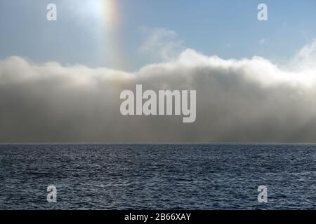 Blick auf den kleinen Regenbogen, der Sonnenhundeeffekt über den Morgennebel genannt wird, der über den Fjord steigt. Stockfoto