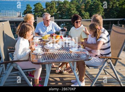 Große glückliche Familie, die zusammen auf der Terrasse frühstücken, am Tisch sitzen und Kaffee trinken. Schöner Meerblick, warmer Sommermorgen. Familie Stockfoto