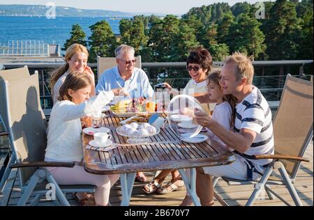 Große glückliche Familie, die zusammen auf der Terrasse frühstücken, am Tisch sitzen und Kaffee trinken. Schöner Meerblick, warmer Sommermorgen. Familie Stockfoto