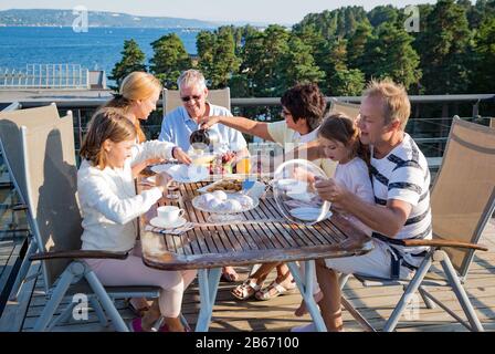 Große glückliche Familie, die zusammen auf der Terrasse frühstücken, am Tisch sitzen und Kaffee trinken. Schöner Meerblick, warmer Sommermorgen. Familie Stockfoto