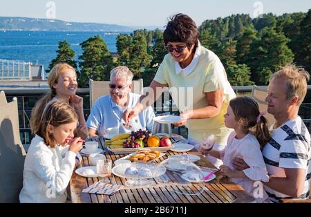 Große glückliche Familie, die zusammen auf der Terrasse frühstücken, am Tisch sitzen und Kaffee trinken. Schöner Meerblick, warmer Sommermorgen. Familie Stockfoto