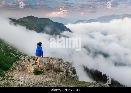 Auf einer Bergkuppe sitzende Touristin mit atemberaubendem Blick auf das nebelige Bergtal. Stockfoto