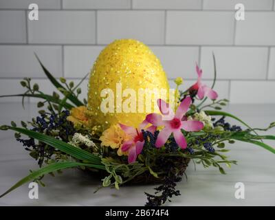 Riesengelbes Osterei schimmernd, in einem Federkranz sitzend mit pinkfarbenen, violetten und gelben Blumen, auf einem Sandsteinfelsen mit einem weißen U-Bahn-Fliesenbein Stockfoto