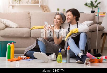 Lächelndes junges Paar, das sich nach der Reinigung des Apartments mit dem Smartphone entspannen kann Stockfoto
