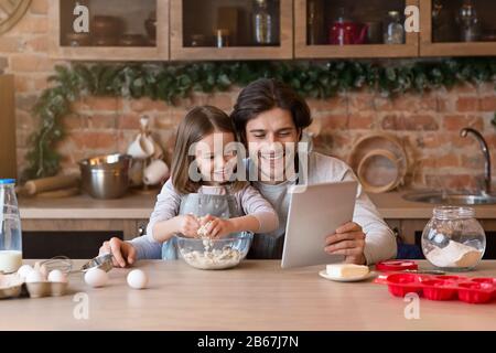 Papa und Tochter machen Teig zusammen und überprüfen das Rezept auf der digitalen Tablette Stockfoto