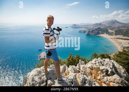 Mann dreht auf der Bergspitze mit der Kamera. Wandern und Fotografieren für Touristen. Panoramablick auf den Strand von Tsampika, Berge und blaues Meer, Rhodes Stockfoto