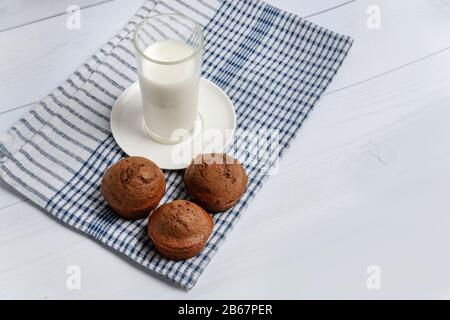 Drei Muffins und ein Glas Milch auf einer Serviette liegen auf einer weißen Holzoberfläche. Stockfoto