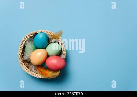 Flache Laienzusammensetzung mit bunten Ostereiern in einem Korb unter Federn auf blauem Grund mit Platz für Text. Stockfoto