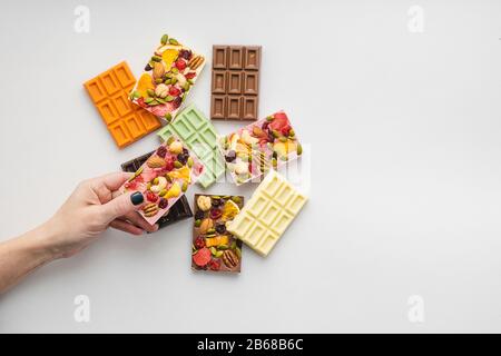 Frau mit heller Maniküre hält in der Hand Schokolade verschiedener Farben und farbige Füllungen mit Scheiben von Fruchtnüssen auf weißem Grund. Oben Stockfoto