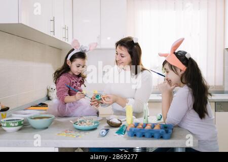 Fröhliche Familie lächelt und lacht, während sie sich auf Ostern vorbereitet. Lächelnde Mutter und Kinder färben Ostereier mit in sonniger Küche. Stockfoto