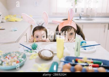 Zwei kleine Mädchen verstecken sich hinter der Küchenecke. Adlige Mädchen in Hünenohren versteckt hinter Küchenbar mit Farbe, Eiern, Bürsten und Osterdekoration Stockfoto