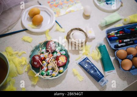 Draufsicht auf die Schüssel mit Eiern und Süßigkeiten für Ostern. Schokoladenmini-Eier in einer Schüssel mit bemalten Eiern auf Küchentisch mit gelben Federn, Gelege, Brushe Stockfoto