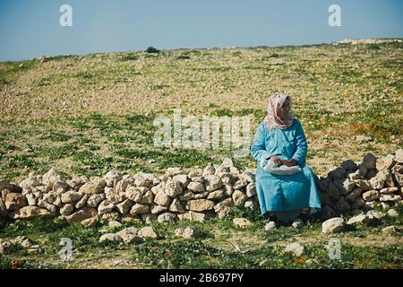 Essaouira, MAROKKO - 19. JANUAR 2020: Arme und einsame marokkanische Frau, die auf Felsen mit Speise- und Wasservorräten sitzt. Stockfoto