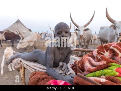 Lächelnder mundarischer Stammjunge, bedeckt mit Asche, die auf einem Bett in einem Viehlager ruht, Central Equatoria, Terekeka, Südsudan Stockfoto