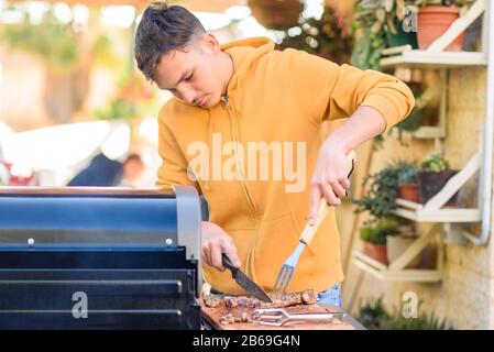 Nahaufnahme des jungen Mannes schneidet Fleisch auf Holzbrett im Freien Picknick. Junger Mann, der Fleisch auf dem Grillgrill auf der Sommerparty im Freien kocht. Stockfoto