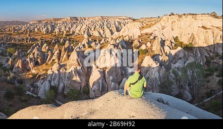 Ein junger Tourist, der auf einer Bergkuppe sitzt und den Sonnenaufgang in Kappadokien im roten Tal betrachtet. Tourismus und Reisen in der Türkei Konzept Stockfoto