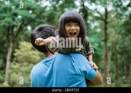 Das kleine Mädchen lachte glücklich, als es während des Spiels im Park auf die Schulter ihres Vaters getragen wurde Stockfoto