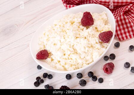 Hüttenkäse mit Beere in weißer Schüssel auf weißem Holztisch. Gesunde Ernährung. Stockfoto