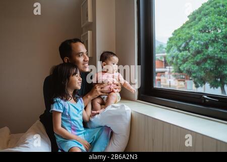 Vater und zwei kleine Tochter, die aus dem Fensterglas sehen, wenn sie im Schlafzimmer auf dem Bett sitzen Stockfoto