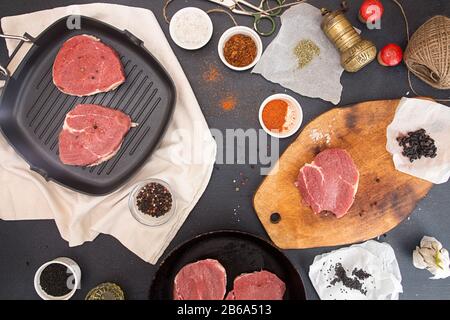 Kochendes Abendessen. Overhead-Stücke roher Schweinesteaks auf Grillpfanne und Hackbrett auf dunklem Hintergrund. Zusammensetzung von Fleisch und Gewürzen. Draufsicht. Stockfoto