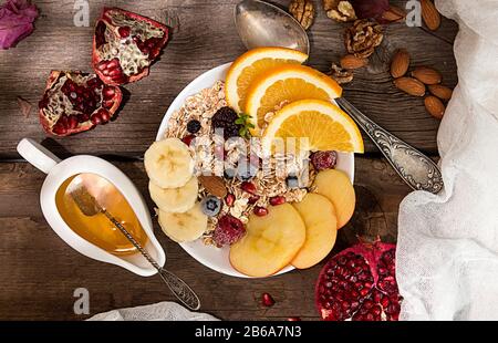 Gesundes Frühstück. Müsli mit orange Frucht, Apfel, Granatapfel, Nüsse, Bananen- und Honig in Weiß Schüssel über auf alten Holztisch. Gesunde organische Natura Stockfoto