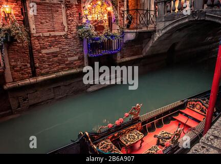 Eine kunstvolle Gondel mit rotem und goldenem Innenraum, die an einem schmalen Kanal in Venedig, Italien, mit Brücke und Weihnachtsbeleuchtung im Hintergrund vermauert ist Stockfoto