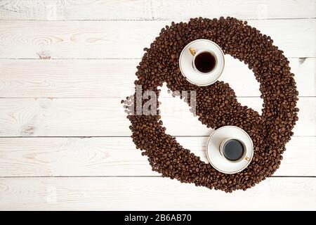 Kaffee-Hintergrund. Haufen Sie geröstete Kaffeebohnen in Form von Yin Yan Zirkel mit Tassen Kaffee auf weißem Hintergrund. Kopierbereich. Draufsicht. Blick von der Abov Stockfoto