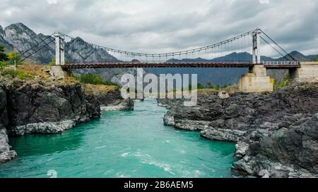 Hängebrücke über einen Gebirgsfluss. Blick vom Fluss. Türkisfarbenes Gebirge, felsige Ufer. Drohnenschießen. Katun River, Altai. Stockfoto