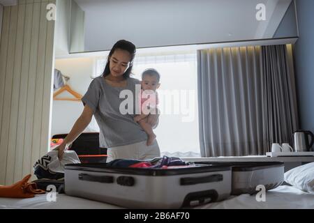 Asiatische Mutter hält ihr Baby bei der Vorbereitung der Kleidung in einen Koffer Stockfoto