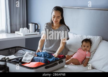 Baby sitzt neben der Mutter und bereitet Kleidung zum Einlegen in den Koffer vor Stockfoto