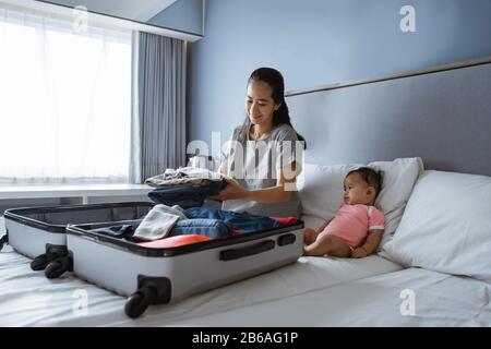 Baby sitzt neben der Mutter und bereitet Kleidung zum Einlegen in den Koffer vor Stockfoto