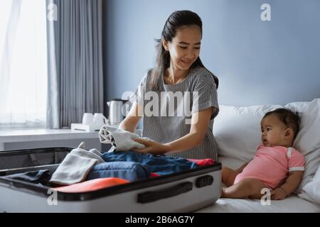 Süßes Baby sitzt neben der Mutter und bereitet Kleidung zum Einlegen in den Koffer vor Stockfoto