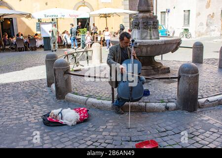 ROM/Italien - 23. April 2019: Ein männlicher Straßenmusiker spielt Cello mit einem kleinen, weißen Hund neben sich und einem Springbrunnen im Hintergrund. Stockfoto