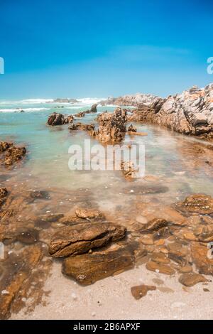 Flaches türkisfarbenes Wasser und Felsen am Strand an einem sonnigen Tag. Der indische Ozean am Kap Agulhas, der südlichsten Spitze Afrikas. Garden Route, Süden Stockfoto