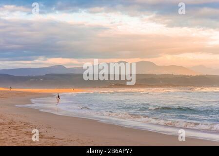 Ein Streifen goldenes Abendlicht, das bei Sonnenuntergang auf den Strand der Plettenberg Bay fällt, mit Bergen in der Ferne und Menschen, die am Strand spazieren gehen. Garde Stockfoto