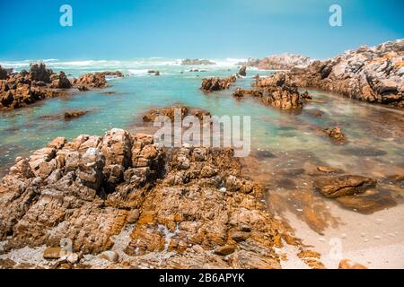 Flaches türkisfarbenes Wasser und Felsen am Strand an einem sonnigen Tag. Der indische Ozean am Kap Agulhas, der südlichsten Spitze Afrikas. Garden Route, Süden Stockfoto