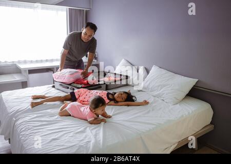 Vater bereitet den Koffer vor, als seine beiden Kinder auf dem Bett im Zimmer spielen Stockfoto