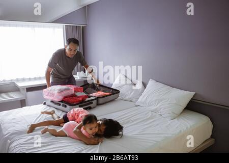 Der asiatische Vater bereitet den Koffer vor, als seine beiden Kinder auf dem Bett im Zimmer spielen Stockfoto