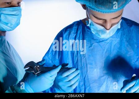 Nahaufnahme, Porträt zweier Chirurgen, in medizinischen Masken und sterilem Gown. Der Assistent gibt den Schlüssel an den Chirurgen weiter. Medizinischer Humor. Chirurgischer Witz. Stockfoto