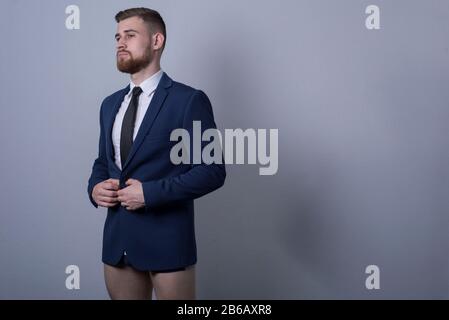 Portrait eines jungen bärtigen, gutaussehenden Geschäftsmannes, in einem Business-Anzug, ohne Hose auf grauem Hintergrund stehend, zur Seite blickend, gerade Stockfoto