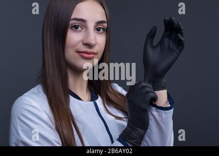 Studio-Porträt eines Arztmädchens, in einem weißen Mantel, lächelnd, mit medizinischen Handschuhen. Auf grauem Hintergrund. Schwarze medizinische Handschuhe. Dramatisches medizinisches PO Stockfoto