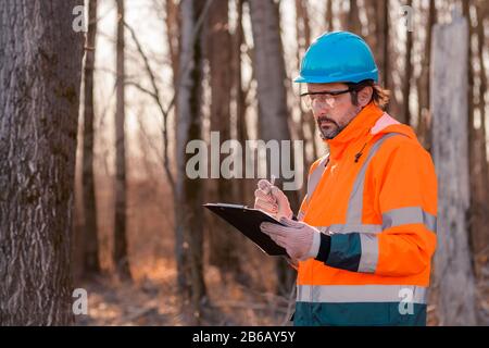 Forsttechniker, der während des Entwaldungsvorgangs Notizen auf Notizblock in der Zwischenablage in Wald schreibt Stockfoto