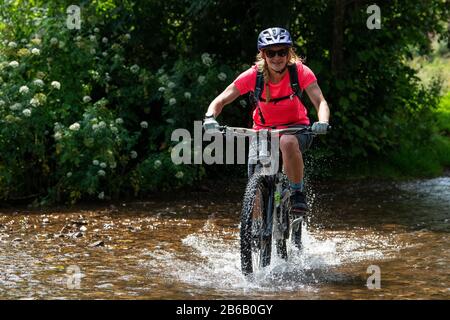 Eine junge blonde Frau fährt mit einem Mountainbike-Rad durch einen Bach in der Nähe von Minehead in Somerset. Stockfoto