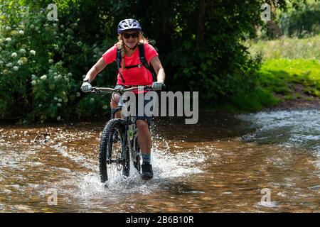 Eine junge blonde Frau fährt mit einem Mountainbike-Rad durch einen Bach in der Nähe von Minehead in Somerset. Stockfoto