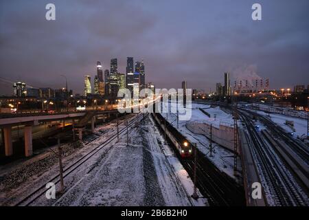 Industrielandschaft mit vielen Bahngleisen weit, Autos auf der Überführung und Wolkenkratzer mit Kraftwerksrohren am Horizont in der Dämmerung im Winter Stockfoto