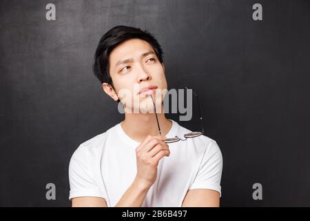 Nahaufnahme des eleganten jungen asiatischen männlichen Studenten, der denkt, die Lippen mit dem Rand der Brille berührt und nachdenkliche, zusammenwirkende Ideen aufsucht, plant Stockfoto