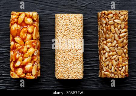 Getreideriegel mit Erdnüssen, Sesam und Sonnenblumenkerne auf einem Schneidebrett auf einem dunklen Steintisch. Ansicht von oben. Drei Verschiedene Balken Stockfoto